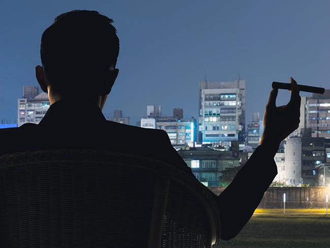 Silhouette of businessman sit on chair and hold a cigar and looking at the city in night.