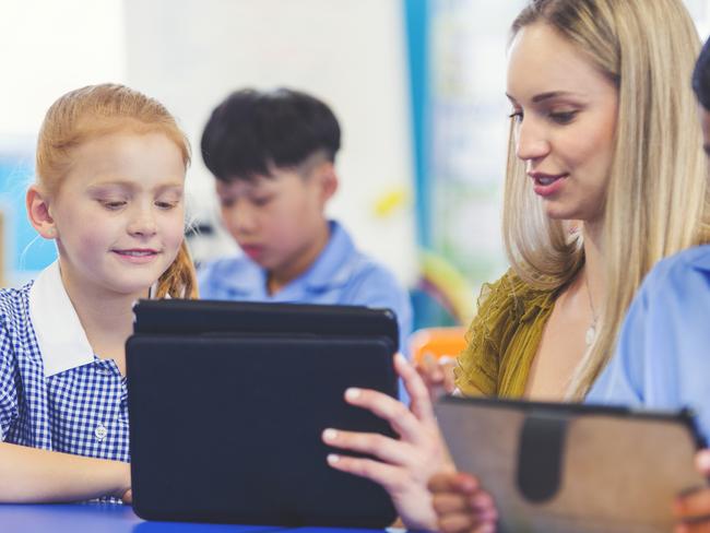 Teacher helping a student in class. They are in a classroom, everyone is wearing a school uniform. They are working at a desk. They are other ethnic backgrounds in shot