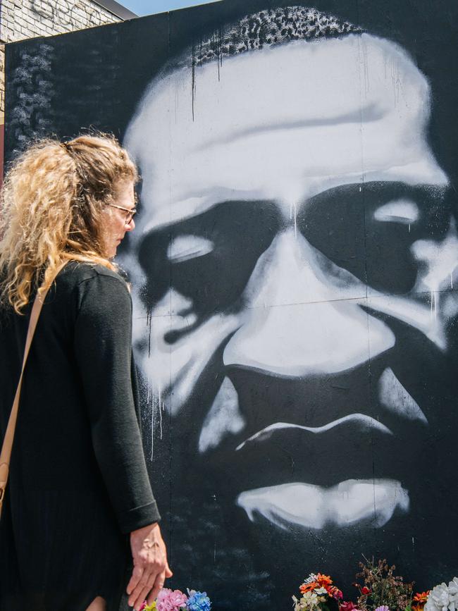 A woman stands in front of a mural of George Floyd.