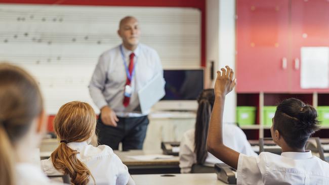 A rear-view shot of teenage music students in a classroom learning how to play the keyboard with their teacher at a school. The students are wearing school uniforms and the teacher is smartly dressed.