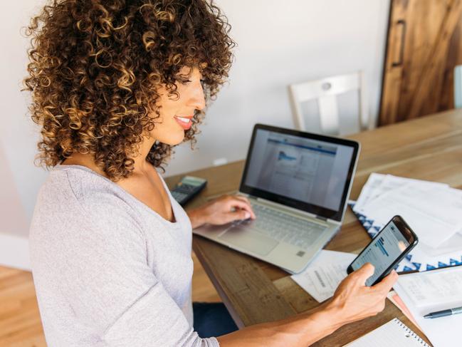 A woman sits at her dining room table with laptop computer and financial reports doing her monthly budget. She is smiling at the ease of use as she works on her smart phone banking app to do monthly finances, pay taxes and save money for the future. She is also filing her tax return. Picture: iStock.