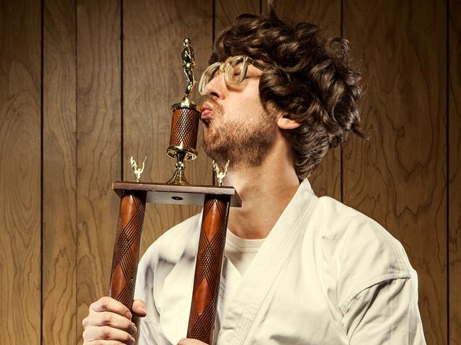 A nerdy looking guy wearing a white gi and black belt with big glasses and a goofy smile holds his new trophy he won at the competition, showing that he has mastered a martial art.   He stands in front of a wood paneled wall kissing the trophy with pursed lips; horizontal with copy space.