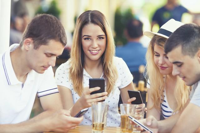 Four teenagers are sitting in a cafe using their mobile phones. Focus on smiling young woman looking at camera