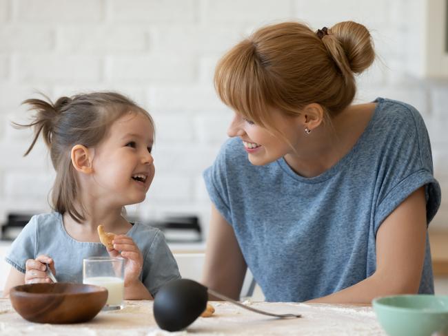 Smiling mother looking at little daughter and feeding her after bake cookies standing at table in kitchen. Happy caring mum and adorable girl eating flour and drink milk, preparing dinner.