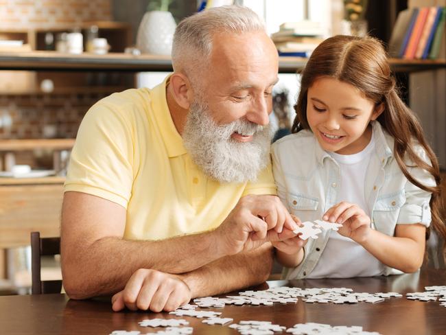 Weekend in harmony. Adorable little girl and her pleasant grandfather sitting at the table, holding pieces of a jigsaw puzzle and trying to connect them together while smiling serenely. Healthy baby boomers and seniors, generic retirement