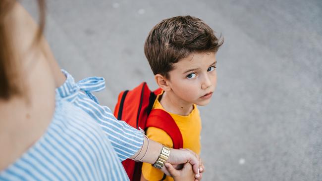 A son with a backpack and his mother holding his hand sending him to school on the first day of school and the schoolboy is looking at the camera