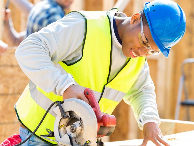 Young African American man concentrates as he uses pwoer tool to prepare wood. He is working with construction crew to build new home. Construction crew is in the background working.