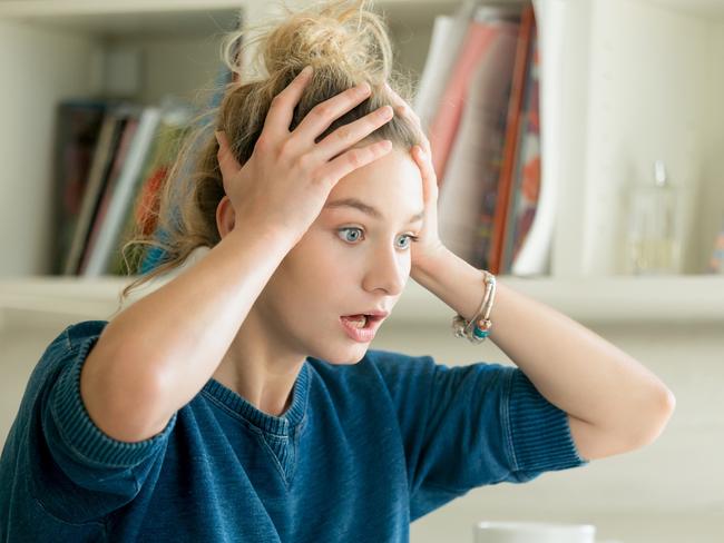 money mistakes - generic woman shocked, stressed... Portrait of an attractive woman at the table with cup and laptop, book, notebook on it, grabbing her head. Bookshelf at the background, concept photo