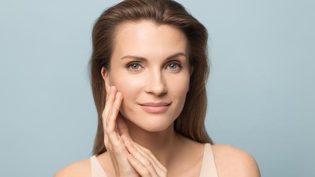 Close up portrait of beautiful young woman isolated on blue studio background touch clean healthy skin, pretty millennial female model look at camera satisfied with moisturised hydrated face treatment