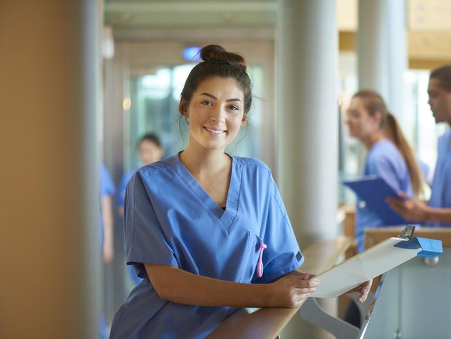 A young female nurse wearing blue scrubs and holding medical records is smiling to camera. She is standing on a busy corridor of a modern hospital . More male and female nurses are walking behind her .