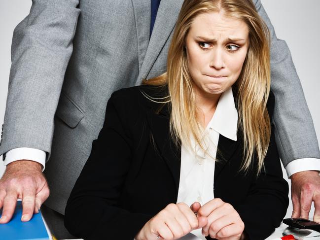 A  businessman stands behind a pretty young businesswoman, arms circling her as she sits at her desk looking threatened, terrified and helpless.