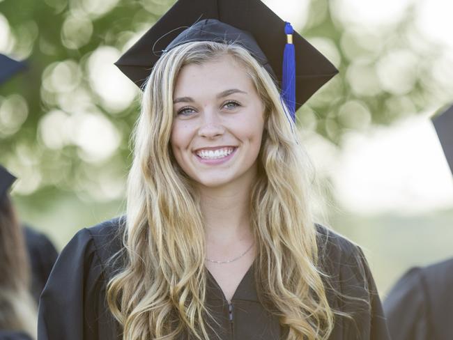 A multi-ethnic group of university students are outdoors at a graduation ceremony. They are wearing graduation robes and caps. They are walking away from the camera, and a Caucasian woman is turned to smile at the camera while holding her diploma.