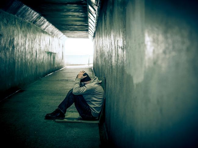 A young homeless male of caucasian ethnicity sits on the floor of a cold, dark subway tunnel, with his hands placed on his head in depair and sadness. He wears jeans, a grey hoodie and a black beanie hat to protect himself from the cold winter weather. He has a beard as well. The image has a heavy vignette and is desaturated to emphasise the bleak subject matter. Horizontal colour image with copy space.