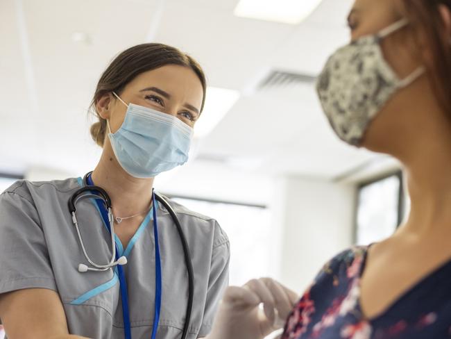 Shot focused on a kind looking nurse wearing scrubs, a stethoscope, white rubber gloves and a protective face mask. She is smiling at the nervous patient with her eyes. The nurse is prepping the patients arm before she injects her with the COVID-19 vaccine.