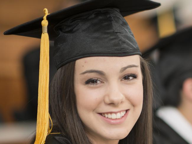 A multi-ethnic group of college graduates are sitting together in a lecture hall and are listening to their professor. They are wearing a cap and gown and one woman is smiling and looking at the camera.