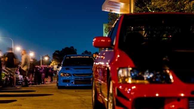 Mitsubishi Evolutions drive down the driveway as people gather around rows of worked cars. Photo: Sam Law - 300C-PO