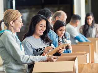 Mature adult Caucasian woman is standing with mid adult Hispanic woman as they pack cardboard boxees full of donated food in charity food bank. Other volunteers are lined up behind them, also sorting donated groceries into boxes. Hispanic woman is writing on checklist on clipboard. Picture: Steve Debenport