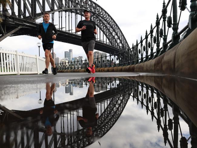 SYDNEY, AUSTRALIA - NewsWire Photos SEPTEMBER 28, 2023: Two morning joggers run past a puddle with Cloudy skies over the Sydney Harbour Bridge reflected in the puddle.Picture: NCA NewsWire / Damian Shaw