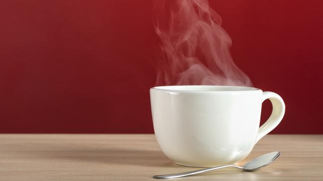 A hot steamy large white cup of coffee or tea with spoon on a blonde colored wood table against a red wall