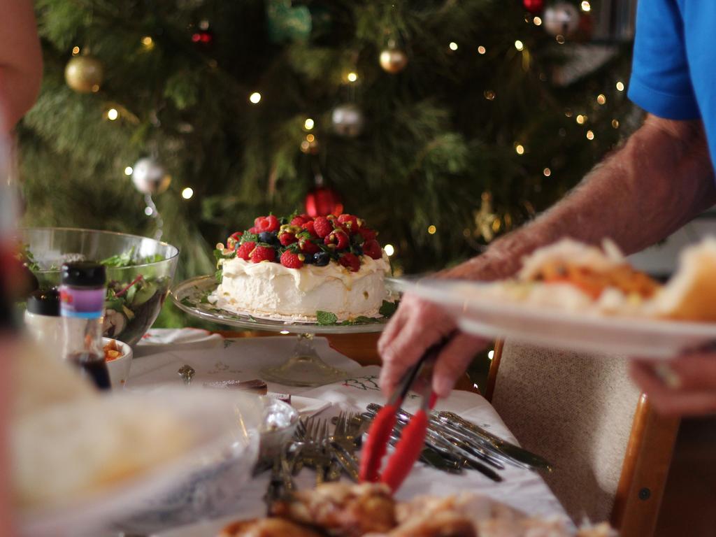 This photo shows a casual Australian Christmas lunch. A Barbecue lunch featuring charcoal grilled chicken, leg of ham, salads and dessert. In the foreground of the photo an elderly man helping himself to the buffet. The mighty pavlova is in focus with its cream, and berry topping. In the background you can see the Christmas tree with fairy lights and decorations.