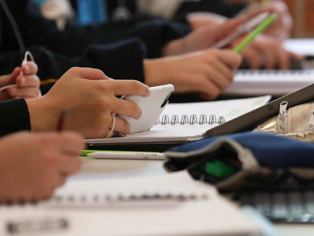 A student in uniform distracted holding using and watching a mobile phone during a lesson at high school. Books, tablets and pencil cases all visible on the desk and work space.