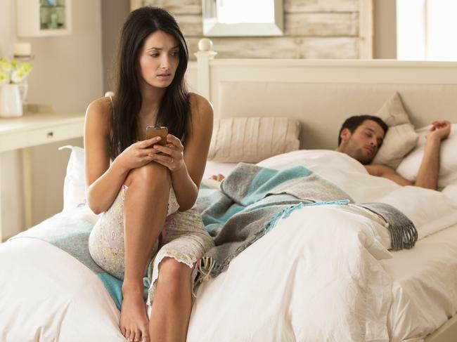 An upset young woman checks her husband's phone as he is sleeping.