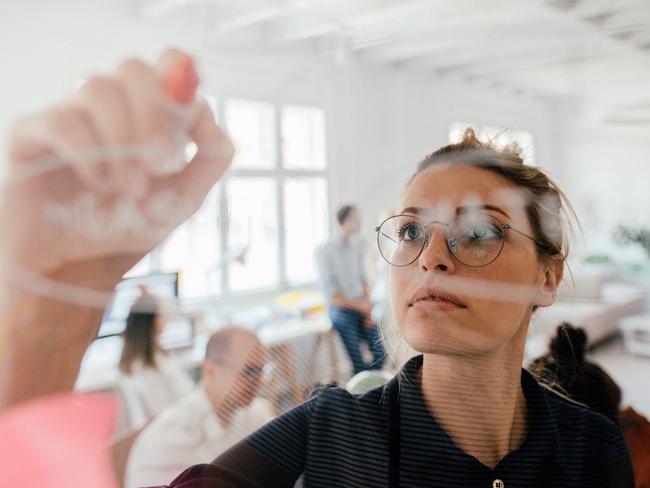 Photo of a young woman writing on a transparent wipe board and thinking of a solution for her work-related problems