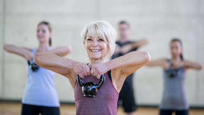 An older Caucasian woman is seen doing a squat with a kettlebell, while participating in a co-ed, multi-ethnic, fitness class. She is expressionless and focusing on her form. Active seniors generic