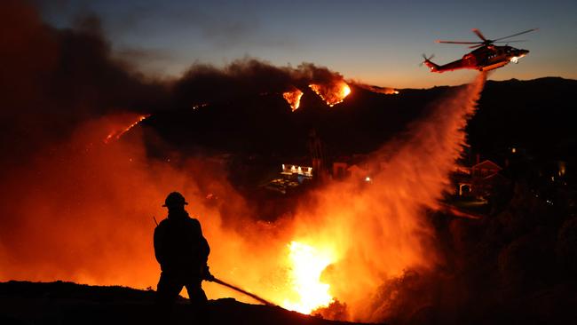 TOPSHOT - Fire personnel respond to homes destroyed while a helicopter drops water as the Palisades Fire grows in Pacific Palisades, California on January 7, 2025. A fast-moving wildfire in a Los Angeles suburb burned buildings and sparked panic, with thousands ordered to evacuate January 7, 2025 as "life threatening" winds whipped the region. Frightened residents abandoned their cars on one of the only roads in and out of the upscale Pacific Palisades area, fleeing on foot from the 770-acre (310-hectare) blaze engulfing an area crammed with multi-million dollar homes in the Santa Monica Mountains. (Photo by David Swanson / AFP)