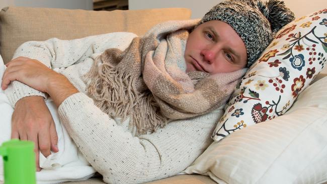 Sick man wearing scarf lying on couch at home under a blanket checking temperature. Living room with table full of medicine and pills. Man with running nose and cough. Istock