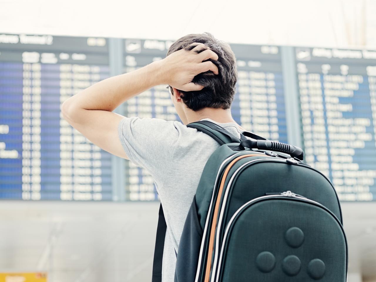 Back view of confused college student traveling with backpack, standing with hand on his head and checking information about his flight on airport timetable - travel concept