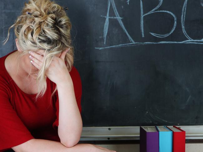 Elementary classroom setting with tired or frustrated teacher holding her head. She's sitting in front of an chalkboard with ABC