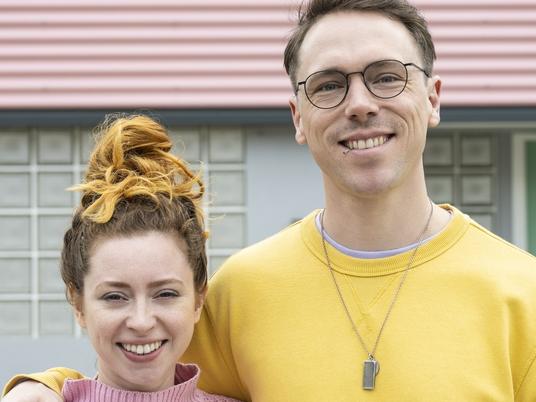 Waist-up view of Caucasian man and woman in casual clothing standing in front of modern style pastel two-story house and smiling at camera.