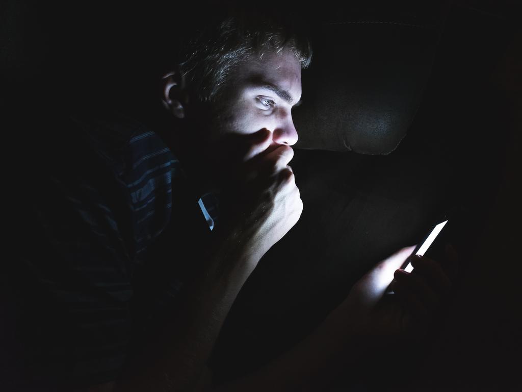 The image displays a teenager reading shocking news on his cell phone as he is lying on a couch in the dark.