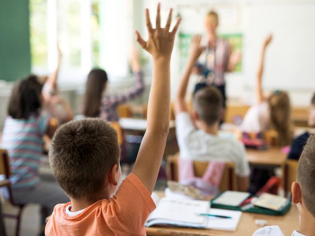 Rear view of little boy and his classmates raising arms to answer teacher's question during the lecture in the classroom.