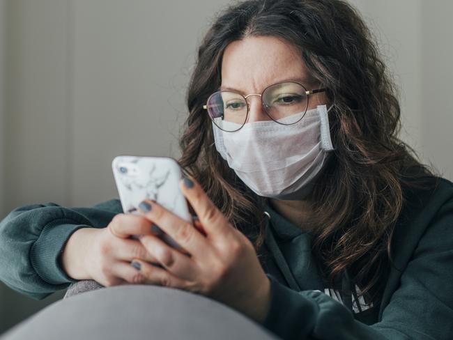 Young woman covering her face with surgical mask and using smartphone to find information about coronavirus spread on internet