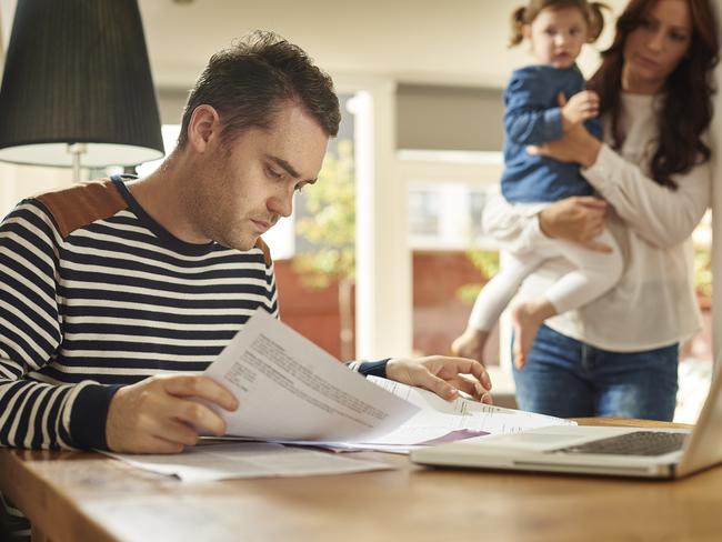 A young husband and father sits at a dining table looking through the household bills and looks worried . His wife and young daughter stand in the background looking on sympathetically .