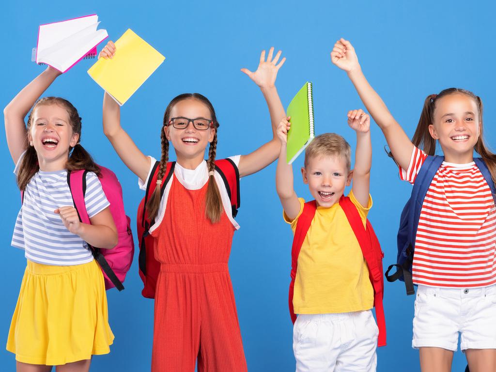 Funny kids classmates with backpacks jumping on blue background from happiness on first school day, raising hands up with exercise books, boys and girls excited to be back at school after vacation