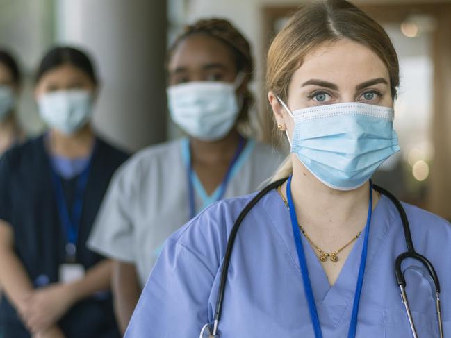 A multi-ethnic team of medical workers wearing scrubs and protective face masks look directly at the camera while posing in a line in a hospital corridor while working during the Covid-19 pandemic. Image is focused on a female doctor standing at the front of the group.