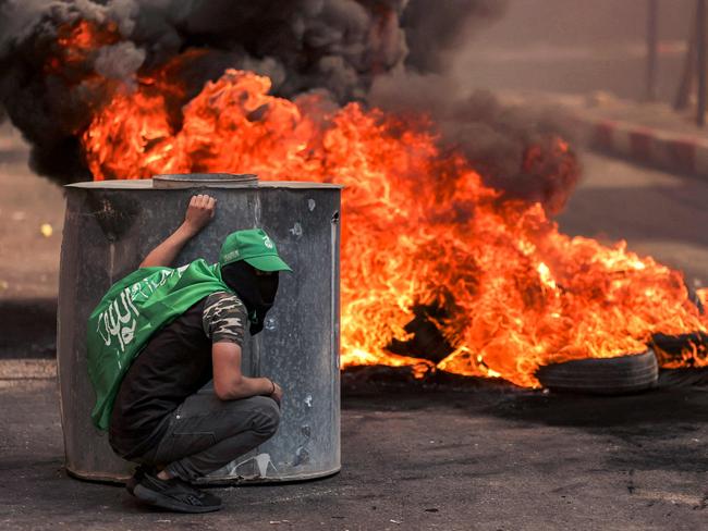 TOPSHOT - A masked Palestinian protester takes cover near flaming tires during clashes with Israeli forces following a rally in solidarity with Gaza by supporters of the Fatah and Hamas movements, in the city of Hebron in the occupied West Bank on October 13, 2023. AFP correspondents and a security official reported clashes after rallies in solidarity with war-battered Gaza in Ramallah, Nablus, Tulkarm, Hebron and other cities and towns, with the Palestinian Red Crescent reporting dozens wounded across the West Bank, some critically. (Photo by HAZEM BADER / AFP)