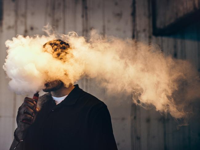 A dark and high contrast image of a young adult male has a cloud of nicotine vapor "smoke" pouring from his mouth.  Vape smoking, or "vaping" is growing in popularity, as well as falling under stricter state and governmental regulations.  Horizontal image with copy space.