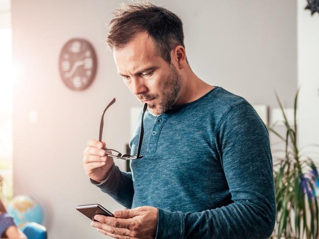 Worried father looking at smart phone with eyeglasses in his hand, in background his daughter doing homework