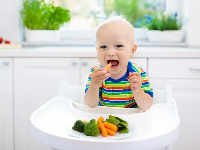 Cute baby eating vegetables in white kitchen. Infant weaning. Little boy trying solid food, organic broccoli, cauliflower, carrot and green peas. Healthy nutrition for kids. Child biting carrot.