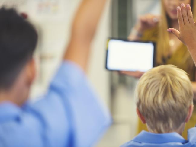 Elementary schoolteacher giving a presentation to the class. The students have their hands raised to ask questions in the classroom