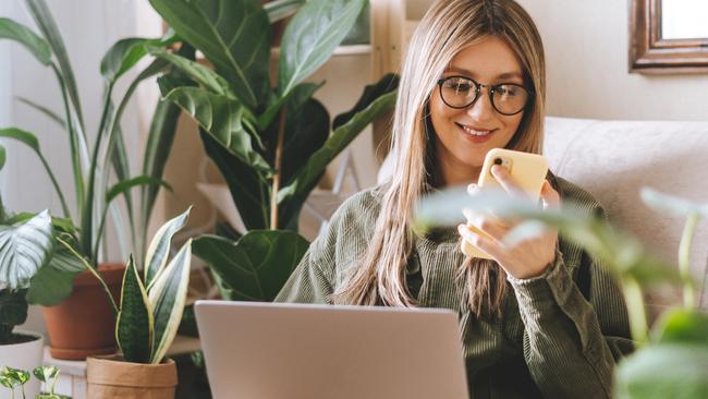 Freelance woman in glasses with mobile phone typing at laptop and working from home office. Happy girl sitting on couch in living room with plants. Distance learning online education and work.