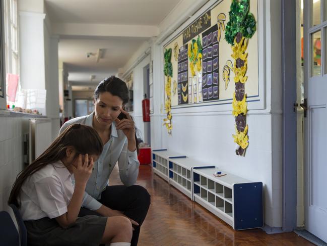 A female teacher sits consoling a young student in the corridor, the little girl looks very upset and holds her head in her hands.