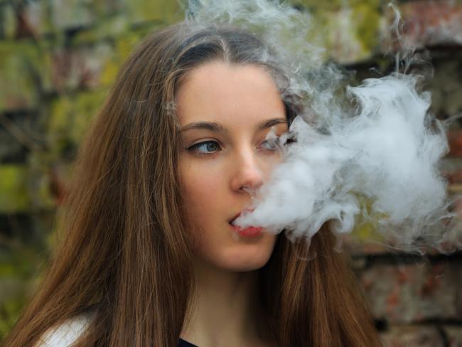 Vape teenager. Young pretty white girl in white cardigan smoking an electronic cigarette opposite destroyed brick wall on the street in the spring. Bad habit that is harmful to health.