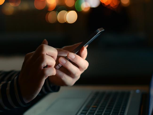 Using smartphone and laptop computer late at night, low key image of freelancer female hands holding mobile phone in dark home office interior and working overtime