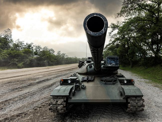 A wide-angle front view of a Japanese self-defense force military tank driving down a dirt road lined with trees with a dramatic sky.