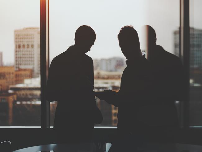 Silhouettes of two businessman near window and table of dark office interior, discussing something and looking on screen of digital tablet, cityscape outside